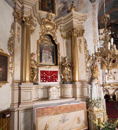 Zdjęcie nr 3: Nastawa ołtarzowa architektoniczna, jednoosiowa, jednokondygnacyjna ze zwieńczeniem, ustawiona na dwustrefowym cokole z wyodrębnionymi prostopadłościanami. Ołtarz kamienny, prostopadłościenny, w drewnianej obudowie. Mensa kamienna z marmurowym portatylem. Antepedium drewniane, w kształcie leżącego prostokąta, ujęte profilowaną ramą, z płaskorzeźbionym monogramem maryjnym oraz fragmentami ornamentu roślinnego. Tabernakulum w kształcie prostopadłościennej skrzynki o wypukłym licu, flankowanej parą kolumienek na cokołach, ustawionych pod kątem, dźwigających woluty oraz gzyms przyjmujący formę łuku segmentowego nadwieszonego. Drzwiczki w kształcie stojącego prostokąta zamkniętego łukiem półkolistym, w opasce, z płaskorzeźbionym przedstawieniem baranka apokaliptycznego, leżącego na księdze, w glorii. Nastawa ujęta dwiema parami pilastrów dekorowanych rocaille'em i stojącą pomiędzy nimi parą kolumn o kapitelach kompozytowych dźwigających pełne belkowanie, wyłamane na ich osi, przerwane pośrodku. Na belkowaniu ustawione dwa wazony. W polu centralnym głęboka wnęka w formie stojącego prostokąta zamkniętego łukiem półkolistym nadwieszonym z uskokiem, ujęta profilowaną ramą, z obrazem Matki Boskiej z Dzieciątkiem w osobnej profilowanej ramie o kształcie stojącego prostokąta zamkniętego ornamentalnym łukiem. Ponad nią, w partii belkowania rokokowy kartusz z rytym monogramem Marii, a poniżej prostokątna płycina z wotami. Pole flankowane dwiema figurami kobiet stojącymi na ćwierćkolistych cokołach. Nastawa ujęta uszami utworzonymi z rocaille'u. Zwieńczenie w formie stojącego prostokąta o wklęsłych bokach, ze spływami z uszakami po bokach, zamknięte gzymsem przyjmującym formę łuku segmentowego nadwieszonego z uskokiem, wyłamanego przy brzegach, dekorowanego rocaille'em. W polu obraz „Nauczanie Marii przez Joachima” ujęty profilowaną ramą w kształcie stojącego prostokąta zamkniętego łukiem segmentowym nadwieszonym z uskokiem. 
Struktura marmoryzowana w kolorze beżowym; ornamentyka, profile i kolumny złocone.