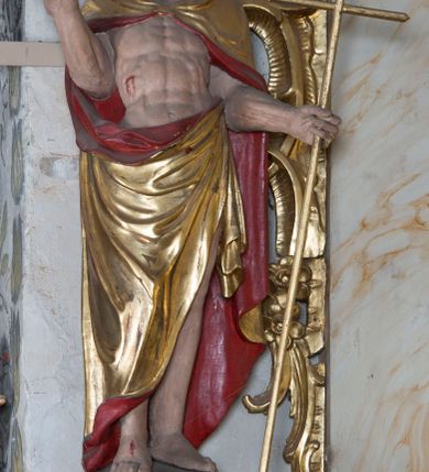 Zdjęcie nr 2: Figura ustawiona na żłobionym cokole na planie kwadratu, o wklęsłych bokach, zwężających się ku górze, dekorowanym perełkowaniem i liśćmi. Chrystus ukazany frontalnie, w wykroku, z głową delikatnie skierowaną w prawo. W lewej, wyciągniętej w bok ręce trzyma laskę zakończoną krzyżem, na której niegdyś zapewne wisiała chorągiew paschalna, prawą unosi w geście błogosławieństwa. Jezus ma pociągłą twarz o wysokim czole, półprzymknięte oczy, długi, prosty nos oraz małe, pełne usta. Twarz okolona jest silnie kręconą, rozdwajającą się brodą oraz kręconymi włosami sięgającymi ramion. Ciało ma szczupłe, silnie umięśnione. Chrystus ubrany jest w złocony płaszcz z czerwoną podszewką zapięty na piersi, zarzucony na ramiona i zawinięty z przodu postaci. Polichromia ciała naturalistyczna, z zaznaczonymi śladami męki. 