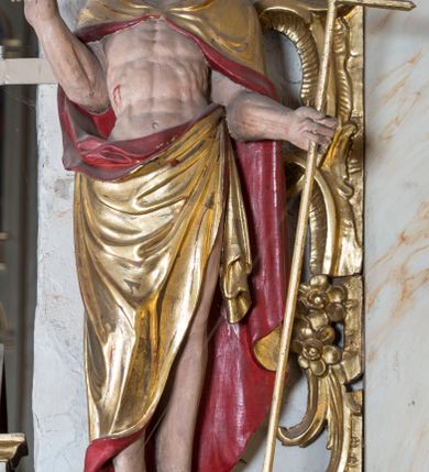 Zdjęcie nr 3: Figura ustawiona na żłobionym cokole na planie kwadratu, o wklęsłych bokach, zwężających się ku górze, dekorowanym perełkowaniem i liśćmi. Chrystus ukazany frontalnie, w wykroku, z głową delikatnie skierowaną w prawo. W lewej, wyciągniętej w bok ręce trzyma laskę zakończoną krzyżem, na której niegdyś zapewne wisiała chorągiew paschalna, prawą unosi w geście błogosławieństwa. Jezus ma pociągłą twarz o wysokim czole, półprzymknięte oczy, długi, prosty nos oraz małe, pełne usta. Twarz okolona jest silnie kręconą, rozdwajającą się brodą oraz kręconymi włosami sięgającymi ramion. Ciało ma szczupłe, silnie umięśnione. Chrystus ubrany jest w złocony płaszcz z czerwoną podszewką zapięty na piersi, zarzucony na ramiona i zawinięty z przodu postaci. Polichromia ciała naturalistyczna, z zaznaczonymi śladami męki. 