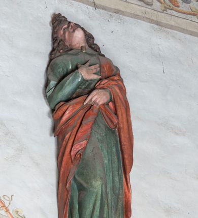 Zdjęcie nr 1: Figura ścięta z tyłu. Święty ukazany frontalnie, w kontrapoście, z głową silnie zadartą do góry, przechyloną w prawo. Lewa ręka podtrzymuje z przodu płaszcz, prawa jest złożona na piersi. Twarz ma szczupłą, o oczach skierowanych w górę, małym nosie i wąskich ustach, okoloną silnie kręconymi włosami sięgającymi ramion, z czubem nad czołem. Święty ubrany jest w przylegającą do ciała zieloną tunikę z kołnierzykiem oraz przewiązany diagonalnie przez lewe ramię czerwony płaszcz. Polichromia naturalistyczna w partiach ciała.
