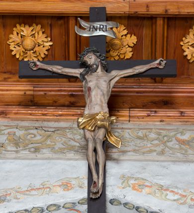 Zdjęcie nr 1: Do prostego krzyża przybita trzema gwoździami figura umarłego Chrystusa. Ciało w lekkim zwisie, o horyzontalnie rozłożonych ramionach, głowie opadającej na prawy bark, lekko ugiętych nogach, stopach skrzyżowanych w układzie prawa na lewą. Twarz ma szczupłą, o zamkniętych oczach, dużym nosie, pełnych ustach, okoloną krótką brodą oraz długimi, kręconymi, ciemnobrązowymi włosami. Na głowie ma koronę cierniową. Ciało wychudzone, o silnie podkreślonym kośćcu, ścięgnach i mięśniach, zwłaszcza w partii klatki piersiowej, z zaznaczonymi śladami męki. Krótkie, złocone perizonium zawinięte wokół podwójnego sznura, ze zwisem tkaniny na lewym boku. Polichromia naturalistyczna w partiach ciała. Pionowa belka krzyża zakończona banderolą z napisem „INRI”. 