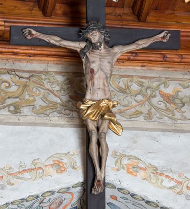 Zdjęcie nr 2: Do prostego krzyża przybita trzema gwoździami figura umarłego Chrystusa. Ciało w lekkim zwisie, o horyzontalnie rozłożonych ramionach, głowie opadającej na prawy bark, lekko ugiętych nogach, stopach skrzyżowanych w układzie prawa na lewą. Twarz ma szczupłą, o zamkniętych oczach, dużym nosie, pełnych ustach, okoloną krótką brodą oraz długimi, kręconymi, ciemnobrązowymi włosami. Na głowie ma koronę cierniową. Ciało wychudzone, o silnie podkreślonym kośćcu, ścięgnach i mięśniach, zwłaszcza w partii klatki piersiowej, z zaznaczonymi śladami męki. Krótkie, złocone perizonium zawinięte wokół podwójnego sznura, ze zwisem tkaniny na lewym boku. Polichromia naturalistyczna w partiach ciała. Pionowa belka krzyża zakończona banderolą z napisem „INRI”. 