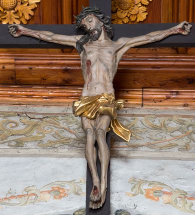 Zdjęcie nr 3: Do prostego krzyża przybita trzema gwoździami figura umarłego Chrystusa. Ciało w lekkim zwisie, o horyzontalnie rozłożonych ramionach, głowie opadającej na prawy bark, lekko ugiętych nogach, stopach skrzyżowanych w układzie prawa na lewą. Twarz ma szczupłą, o zamkniętych oczach, dużym nosie, pełnych ustach, okoloną krótką brodą oraz długimi, kręconymi, ciemnobrązowymi włosami. Na głowie ma koronę cierniową. Ciało wychudzone, o silnie podkreślonym kośćcu, ścięgnach i mięśniach, zwłaszcza w partii klatki piersiowej, z zaznaczonymi śladami męki. Krótkie, złocone perizonium zawinięte wokół podwójnego sznura, ze zwisem tkaniny na lewym boku. Polichromia naturalistyczna w partiach ciała. Pionowa belka krzyża zakończona banderolą z napisem „INRI”. 