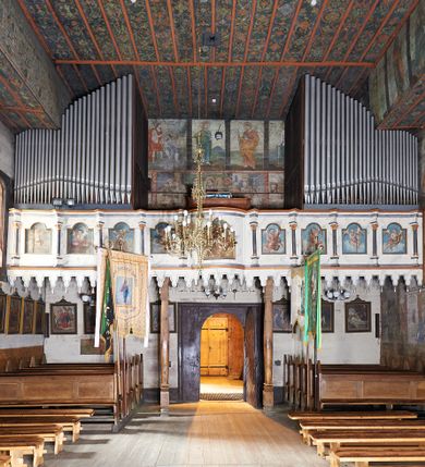 Zdjęcie nr 1: Balustrada chóru muzycznego dziewięcioosiowa, jednokondygnacyjna, od spodu z podwieszonym lambrekinem, na osi środkowej wypukła. Osie flankowane kolumienkami ustawionymi na cokołach, które podtrzymują imposty ustawione pod kątem, wyłamujące profilowany gzyms balustrady. Trzony kolumienek malowane na czarno, kapitele stylizowane na korynckie, złocone. W każdej osi umieszczona płycina w kształcie stojącego prostokąta zamknięta łukiem nadwieszonym. Kolejno od lewej strony malowane w płycinach obrazy: aniołek grający na trąbce, aniołek grający na bębnach, ponownie aniołek grający na trąbce i aniołek grający na skrzypach. Na środku osi płycina w kształcie stojącego prostokąta zamknięta trójkątnie o wyłamanym zwieńczeniu, w polu przedstawienie króla Dawida grającego na harfie. U góry banderola z napisem „LAUDATE DOMINUM IN CITHARA”. Po drugiej stronie balustrady, kolejno od lewej: aniołek grający na wiolonczeli, aniołek grający na trąbce, aniołek grający na flecie i aniołek grający na mandolinie. Wszystkie postacie ukazane na jasnobłękitnym tle, na chmurze białych obłoków. Anioły półnagie, ubrane w czerwone szaty. Struktura balustrady marmoryzowana na biało z niebieskimi przetarciami, detale złocone i malowane na czarno.
