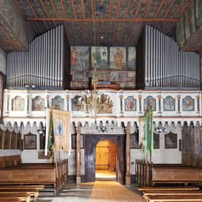 Zdjęcie nr 1: Balustrada chóru muzycznego dziewięcioosiowa, jednokondygnacyjna, od spodu z podwieszonym lambrekinem, na osi środkowej wypukła. Osie flankowane kolumienkami ustawionymi na cokołach, które podtrzymują imposty ustawione pod kątem, wyłamujące profilowany gzyms balustrady. Trzony kolumienek malowane na czarno, kapitele stylizowane na korynckie, złocone. W każdej osi umieszczona płycina w kształcie stojącego prostokąta zamknięta łukiem nadwieszonym. Kolejno od lewej strony malowane w płycinach obrazy: aniołek grający na trąbce, aniołek grający na bębnach, ponownie aniołek grający na trąbce i aniołek grający na skrzypach. Na środku osi płycina w kształcie stojącego prostokąta zamknięta trójkątnie o wyłamanym zwieńczeniu, w polu przedstawienie króla Dawida grającego na harfie. U góry banderola z napisem „LAUDATE DOMINUM IN CITHARA”. Po drugiej stronie balustrady, kolejno od lewej: aniołek grający na wiolonczeli, aniołek grający na trąbce, aniołek grający na flecie i aniołek grający na mandolinie. Wszystkie postacie ukazane na jasnobłękitnym tle, na chmurze białych obłoków. Anioły półnagie, ubrane w czerwone szaty. Struktura balustrady marmoryzowana na biało z niebieskimi przetarciami, detale złocone i malowane na czarno.
