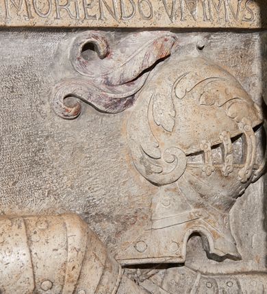 Zdjęcie nr 5: Płyta nagrobna z płaskorzeźbionym przedstawieniem rycerza w typie sansovinowskim, ukazanym w półśnie. Rycerz ułożony na prawym boku, prawą ręką podpierający głowę spoczywającą na poduszce, z lewą złożoną na pancerzu; nogi zgięte w kolanach i skrzyżowane. W prawym górnym narożu płyty umieszczony hełm z opuszczoną zasłoną prętową i pióropuszem. Rycerz ubrany w zbroję turniejową, z przypiętą do pasa po prawej stronie szablą, batorówką i ostrogami na butach. Włosy ułożone w stylu polskim, ma wąsy. Wzdłuż krawędzi prawego boku na kamieniu płyty nagrobnej znajduje się inskrypcja. Bezpośrednio ponad głową zmarłego napis „VIVIENDO MOBIMVR”, zaś nad hełmem „MORIENDO VIVIMVS”. Nad płytą nagrobną barokowy kartusz z tarczą herbową. Tarcza owalna, wypukła, czteropolowa, ujęta ornamentem rollwerkowym. W skwadrowanym polu tarczy umieszczono zatarte godła herbów: Rawicz (pole I), Topór (pole II), Rogala (pole III), Półkozic (pole IV).
