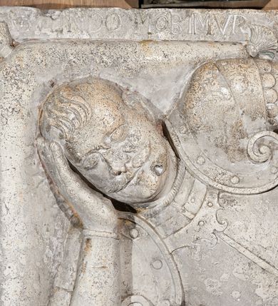 Zdjęcie nr 2: Płyta nagrobna z płaskorzeźbionym przedstawieniem rycerza w typie sansovinowskim, ukazanym w półśnie. Rycerz ułożony na prawym boku, prawą ręką podpierający głowę spoczywającą na poduszce, z lewą złożoną na pancerzu; nogi zgięte w kolanach i skrzyżowane. W prawym górnym narożu płyty umieszczony hełm z opuszczoną zasłoną prętową i pióropuszem. Rycerz ubrany w zbroję turniejową, z przypiętą do pasa po prawej stronie szablą, batorówką i ostrogami na butach. Włosy ułożone w stylu polskim, ma wąsy. Wzdłuż krawędzi prawego boku na kamieniu płyty nagrobnej znajduje się inskrypcja. Bezpośrednio ponad głową zmarłego napis „VIVIENDO MOBIMVR”, zaś nad hełmem „MORIENDO VIVIMVS”. Nad płytą nagrobną barokowy kartusz z tarczą herbową. Tarcza owalna, wypukła, czteropolowa, ujęta ornamentem rollwerkowym. W skwadrowanym polu tarczy umieszczono zatarte godła herbów: Rawicz (pole I), Topór (pole II), Rogala (pole III), Półkozic (pole IV).
