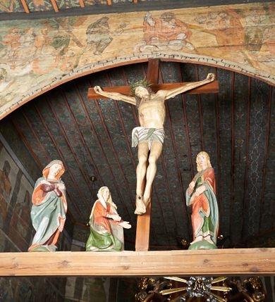 Zdjęcie nr 1: Łuk tęczowy o półkolistym wykroju wsparty na kroksztynach. Belka profilowana, współczesna (z czasów ostatniej konserwacji, nawiązująca do poprzedniej gotyckiej). W centrum krucyfiks z postacią Chrystusa umarłego, po prawej św. Jan Ewangelista, po lewej Matka Boska i św. Maria Magdalena. Chrystus przybity do krzyża trzema gwoździami, sylwetka w delikatnym zwisie, z głową przechyloną na prawe ramię, o silnie podkreślonej anatomii ciała, z zaznaczonymi śladami męki. Perizonium w formie tkaniny przewiązanej na prawym biodrze. Twarz podłużna, okolona zarostem; włosy długie spływające na prawe ramię w kręcone pukle, na głowie korona cierniowa. Po prawej stronie rzeźba św. Jana Ewangelisty, ukazanego w postawie stojącej, zwróconego w trzech czwartych w prawo, lewą ręką przytrzymującego z przodu płaszcz. Święty jest ubrany w długą, zieloną suknię z długimi rękawami oraz czerwony płaszcz, spięty na piersi, zdobiony na brzegach grubą lamelką. Twarz owalna, policzki silnie zarumienione, małe usta, długi i wąski nos, wzrok skierowany przed siebie, zmarszczone brwi. Włosy jasne, spływające do ramion ciasno ułożonymi puklami. Po lewej stronie krucyfiksu rzeźba św. Marii Magdaleny ukazana w pozycji klęczącej, z prawego profilu, z wyciągniętymi w stronę krzyża dłońmi. Święta jest ubrana w długą, czerwoną suknię oraz obszerny zielony płaszcz ze srebrną podszewką, na głowie ma biały welon. Twarz owalna, z wyraźnie zaznaczonym podbródkiem, zmarszczone brwi; spod welonu spływają na lewe ramię długie i jasne włosy. Za nią rzeźba Matki Boskiej ustawiona w kontrapoście, z prawą nogą ugiętą w kolanie, ze skrzyżowanymi na piersi rękami. Maria jest ubrana w długą, bordową suknię oraz błękitny płaszcz z czerwoną podszewką. Twarz szeroka, z wyraźnie zaznaczonym podbródkiem, wzrok skierowany w dal, zmarszczone brwi. Rzeźby ustawione na półkolistych, zielonych cokolikach. Polichromia w odsłoniętych partiach ciała naturalistyczna, detale złocone. 
