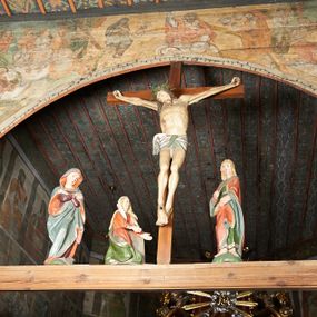 Zdjęcie nr 1: Łuk tęczowy o półkolistym wykroju wsparty na kroksztynach. Belka profilowana, współczesna (z czasów ostatniej konserwacji, nawiązująca do poprzedniej gotyckiej). W centrum krucyfiks z postacią Chrystusa umarłego, po prawej św. Jan Ewangelista, po lewej Matka Boska i św. Maria Magdalena. Chrystus przybity do krzyża trzema gwoździami, sylwetka w delikatnym zwisie, z głową przechyloną na prawe ramię, o silnie podkreślonej anatomii ciała, z zaznaczonymi śladami męki. Perizonium w formie tkaniny przewiązanej na prawym biodrze. Twarz podłużna, okolona zarostem; włosy długie spływające na prawe ramię w kręcone pukle, na głowie korona cierniowa. Po prawej stronie rzeźba św. Jana Ewangelisty, ukazanego w postawie stojącej, zwróconego w trzech czwartych w prawo, lewą ręką przytrzymującego z przodu płaszcz. Święty jest ubrany w długą, zieloną suknię z długimi rękawami oraz czerwony płaszcz, spięty na piersi, zdobiony na brzegach grubą lamelką. Twarz owalna, policzki silnie zarumienione, małe usta, długi i wąski nos, wzrok skierowany przed siebie, zmarszczone brwi. Włosy jasne, spływające do ramion ciasno ułożonymi puklami. Po lewej stronie krucyfiksu rzeźba św. Marii Magdaleny ukazana w pozycji klęczącej, z prawego profilu, z wyciągniętymi w stronę krzyża dłońmi. Święta jest ubrana w długą, czerwoną suknię oraz obszerny zielony płaszcz ze srebrną podszewką, na głowie ma biały welon. Twarz owalna, z wyraźnie zaznaczonym podbródkiem, zmarszczone brwi; spod welonu spływają na lewe ramię długie i jasne włosy. Za nią rzeźba Matki Boskiej ustawiona w kontrapoście, z prawą nogą ugiętą w kolanie, ze skrzyżowanymi na piersi rękami. Maria jest ubrana w długą, bordową suknię oraz błękitny płaszcz z czerwoną podszewką. Twarz szeroka, z wyraźnie zaznaczonym podbródkiem, wzrok skierowany w dal, zmarszczone brwi. Rzeźby ustawione na półkolistych, zielonych cokolikach. Polichromia w odsłoniętych partiach ciała naturalistyczna, detale złocone. 
