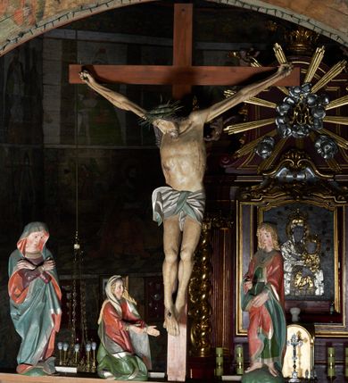 Zdjęcie nr 6: Łuk tęczowy o półkolistym wykroju wsparty na kroksztynach. Belka profilowana, współczesna (z czasów ostatniej konserwacji, nawiązująca do poprzedniej gotyckiej). W centrum krucyfiks z postacią Chrystusa umarłego, po prawej św. Jan Ewangelista, po lewej Matka Boska i św. Maria Magdalena. Chrystus przybity do krzyża trzema gwoździami, sylwetka w delikatnym zwisie, z głową przechyloną na prawe ramię, o silnie podkreślonej anatomii ciała, z zaznaczonymi śladami męki. Perizonium w formie tkaniny przewiązanej na prawym biodrze. Twarz podłużna, okolona zarostem; włosy długie spływające na prawe ramię w kręcone pukle, na głowie korona cierniowa. Po prawej stronie rzeźba św. Jana Ewangelisty, ukazanego w postawie stojącej, zwróconego w trzech czwartych w prawo, lewą ręką przytrzymującego z przodu płaszcz. Święty jest ubrany w długą, zieloną suknię z długimi rękawami oraz czerwony płaszcz, spięty na piersi, zdobiony na brzegach grubą lamelką. Twarz owalna, policzki silnie zarumienione, małe usta, długi i wąski nos, wzrok skierowany przed siebie, zmarszczone brwi. Włosy jasne, spływające do ramion ciasno ułożonymi puklami. Po lewej stronie krucyfiksu rzeźba św. Marii Magdaleny ukazana w pozycji klęczącej, z prawego profilu, z wyciągniętymi w stronę krzyża dłońmi. Święta jest ubrana w długą, czerwoną suknię oraz obszerny zielony płaszcz ze srebrną podszewką, na głowie ma biały welon. Twarz owalna, z wyraźnie zaznaczonym podbródkiem, zmarszczone brwi; spod welonu spływają na lewe ramię długie i jasne włosy. Za nią rzeźba Matki Boskiej ustawiona w kontrapoście, z prawą nogą ugiętą w kolanie, ze skrzyżowanymi na piersi rękami. Maria jest ubrana w długą, bordową suknię oraz błękitny płaszcz z czerwoną podszewką. Twarz szeroka, z wyraźnie zaznaczonym podbródkiem, wzrok skierowany w dal, zmarszczone brwi. Rzeźby ustawione na półkolistych, zielonych cokolikach. Polichromia w odsłoniętych partiach ciała naturalistyczna, detale złocone. 
