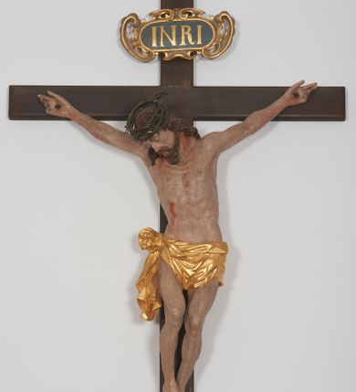 Zdjęcie nr 1: Krucyfiks z rzeźbą Chrystusa w typie Cristo morte. Rzeźba pełna; ciało w dużym zwisie, o esowato wygiętej sylwetce; głowa przechylona na prawe ramię; nogi ugięte w kolanach, stopy przebite jednym gwoździem w układzie prawa na lewą. Twarz pociągła, o wyraźnych rysach, okolona krótkim zarostem. Na głowie szeroka korona cierniowa. Sylwetka Chrystusa wychudzona, ciało z zaznaczonymi żebrami klatki piersiowej i mostkiem. Perizonium przewiązane na prawym boku. Nad głową Chrystusa znajduje się owalny kartusz obwiedziony ażurowym ornamentem małżowinowo-chrząstkowym z napisem „INRI” w polu. Polichromia naturalistyczna w odsłoniętych partiach ciała z zaznaczonymi śladami męki, perizonium złocone.