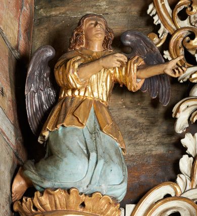 Zdjęcie nr 2: Rzeźby aniołów flankujące cokół nastawy ołtarzowej ukazane są w pozycji stojącej, zwrócone przeciwstawnie, w trzech czwartych ku środkowi nastawy z wyciągniętymi doń rękami. Ubrane w długie zielone suknie, z rozcięciem odsłaniającym jedną nogę powyżej kolana oraz złoconą tunikę z podwiniętymi rękawami do łokci i kołnierzykiem. Anioły mają po jednym skrzydle, usytuowanym od strony zewnętrznej. Modelunek postaci jest sztywny. Anioły osadzone po bokach zwieńczenia ukazane są w pozycji klęczącej, przeciwstawnie do siebie, zwrócone w trzech czwartych ku środkowi, z wyciągniętymi doń rękami. Ubrane w długie jasnozielone suknie oraz złocone tuniki z podwiniętymi rękawami do łokci i kołnierzykiem. Anioły mają dwa skrzydła, szeroko rozłożone. Anioł u szczytu nastawy jest ukazany w pozycji stojącej, frontalnie, w delikatnym kontrapoście, z rozłożonymi na boki rękami, z głową zwróconą w prawo. Ubrany analogicznie jak anioły flankujące zwieńczenie. 
Twarze wszystkich aniołów owalne, z silnie wysuniętym podbródkiem, zarumienionymi policzkami i małymi oczami. Włosy długie, ciemnobrązowe, kędzierzawe, z przedziałkiem pośrodku, opadające na plecy. Polichromia w odsłoniętych partiach ciała naturalistyczna, skrzydła srebrzone. 
