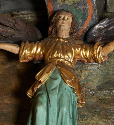 Zdjęcie nr 1: Rzeźby aniołów flankujące cokół nastawy ołtarzowej ukazane są w pozycji stojącej, zwrócone przeciwstawnie, w trzech czwartych ku środkowi nastawy z wyciągniętymi doń rękami. Ubrane w długie zielone suknie, z rozcięciem odsłaniającym jedną nogę powyżej kolana oraz złoconą tunikę z podwiniętymi rękawami do łokci i kołnierzykiem. Anioły mają po jednym skrzydle, usytuowanym od strony zewnętrznej. Modelunek postaci jest sztywny. Anioły osadzone po bokach zwieńczenia ukazane są w pozycji klęczącej, przeciwstawnie do siebie, zwrócone w trzech czwartych ku środkowi, z wyciągniętymi doń rękami. Ubrane w długie jasnozielone suknie oraz złocone tuniki z podwiniętymi rękawami do łokci i kołnierzykiem. Anioły mają dwa skrzydła, szeroko rozłożone. Anioł u szczytu nastawy jest ukazany w pozycji stojącej, frontalnie, w delikatnym kontrapoście, z rozłożonymi na boki rękami, z głową zwróconą w prawo. Ubrany analogicznie jak anioły flankujące zwieńczenie. 
Twarze wszystkich aniołów owalne, z silnie wysuniętym podbródkiem, zarumienionymi policzkami i małymi oczami. Włosy długie, ciemnobrązowe, kędzierzawe, z przedziałkiem pośrodku, opadające na plecy. Polichromia w odsłoniętych partiach ciała naturalistyczna, skrzydła srebrzone. 
