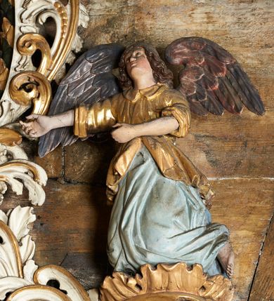 Zdjęcie nr 5: Rzeźby aniołów flankujące cokół nastawy ołtarzowej ukazane są w pozycji stojącej, zwrócone przeciwstawnie, w trzech czwartych ku środkowi nastawy z wyciągniętymi doń rękami. Ubrane w długie zielone suknie, z rozcięciem odsłaniającym jedną nogę powyżej kolana oraz złoconą tunikę z podwiniętymi rękawami do łokci i kołnierzykiem. Anioły mają po jednym skrzydle, usytuowanym od strony zewnętrznej. Modelunek postaci jest sztywny. Anioły osadzone po bokach zwieńczenia ukazane są w pozycji klęczącej, przeciwstawnie do siebie, zwrócone w trzech czwartych ku środkowi, z wyciągniętymi doń rękami. Ubrane w długie jasnozielone suknie oraz złocone tuniki z podwiniętymi rękawami do łokci i kołnierzykiem. Anioły mają dwa skrzydła, szeroko rozłożone. Anioł u szczytu nastawy jest ukazany w pozycji stojącej, frontalnie, w delikatnym kontrapoście, z rozłożonymi na boki rękami, z głową zwróconą w prawo. Ubrany analogicznie jak anioły flankujące zwieńczenie. 
Twarze wszystkich aniołów owalne, z silnie wysuniętym podbródkiem, zarumienionymi policzkami i małymi oczami. Włosy długie, ciemnobrązowe, kędzierzawe, z przedziałkiem pośrodku, opadające na plecy. Polichromia w odsłoniętych partiach ciała naturalistyczna, skrzydła srebrzone. 
