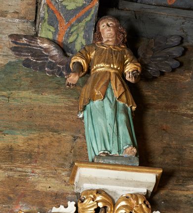 Zdjęcie nr 8: Rzeźby aniołów flankujące cokół nastawy ołtarzowej ukazane w pozycji stojącej, zwrócone przeciwstawnie, w trzech czwartych ku środkowi nastawy z wyciągniętymi doń rękami. Ubrane w długie zielone suknie, z rozcięciem odsłaniającym jedną nogę powyżej kolana oraz złoconą tunikę z podwiniętymi rękawami do łokci i kołnierzykiem. Anioły mają po jednym skrzydle, usytuowanym od strony zewnętrznej. Modelunek postaci jest sztywny. Anioły osadzone po bokach zwieńczenia ukazane są w pozycji klęczącej, przeciwstawnie do siebie, zwrócone w trzech czwartych ku środkowi, z wyciągniętymi doń rękami. Ubrane w długie jasnozielone suknie oraz złocone tuniki z podwiniętymi rękawami do łokci i kołnierzykiem. Anioły mają dwa skrzydła, szeroko rozłożone. Anioł u szczytu nastawy jest ukazany w pozycji stojącej, frontalnie, w delikatnym kontrapoście, z wyciągniętymi do przodu rękami, z głową zwróconą w prawo, z rozłożonymi szeroko skrzydłami. Ubrany analogicznie jak anioły flankujące zwieńczenie. 
Twarze wszystkich aniołów owalne, z silnie wysuniętym podbródkiem, zarumienionymi policzkami i małymi oczami. Włosy długie, ciemnobrązowe, kędzierzawe, z przedziałkiem pośrodku, opadające na plecy. Polichromia w odsłoniętych partiach ciała naturalistyczna, skrzydła srebrzone. 
