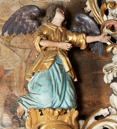 Zdjęcie nr 9: Rzeźby aniołów flankujące cokół nastawy ołtarzowej ukazane w pozycji stojącej, zwrócone przeciwstawnie, w trzech czwartych ku środkowi nastawy z wyciągniętymi doń rękami. Ubrane w długie zielone suknie, z rozcięciem odsłaniającym jedną nogę powyżej kolana oraz złoconą tunikę z podwiniętymi rękawami do łokci i kołnierzykiem. Anioły mają po jednym skrzydle, usytuowanym od strony zewnętrznej. Modelunek postaci jest sztywny. Anioły osadzone po bokach zwieńczenia ukazane są w pozycji klęczącej, przeciwstawnie do siebie, zwrócone w trzech czwartych ku środkowi, z wyciągniętymi doń rękami. Ubrane w długie jasnozielone suknie oraz złocone tuniki z podwiniętymi rękawami do łokci i kołnierzykiem. Anioły mają dwa skrzydła, szeroko rozłożone. Anioł u szczytu nastawy jest ukazany w pozycji stojącej, frontalnie, w delikatnym kontrapoście, z wyciągniętymi do przodu rękami, z głową zwróconą w prawo, z rozłożonymi szeroko skrzydłami. Ubrany analogicznie jak anioły flankujące zwieńczenie. 
Twarze wszystkich aniołów owalne, z silnie wysuniętym podbródkiem, zarumienionymi policzkami i małymi oczami. Włosy długie, ciemnobrązowe, kędzierzawe, z przedziałkiem pośrodku, opadające na plecy. Polichromia w odsłoniętych partiach ciała naturalistyczna, skrzydła srebrzone. 
