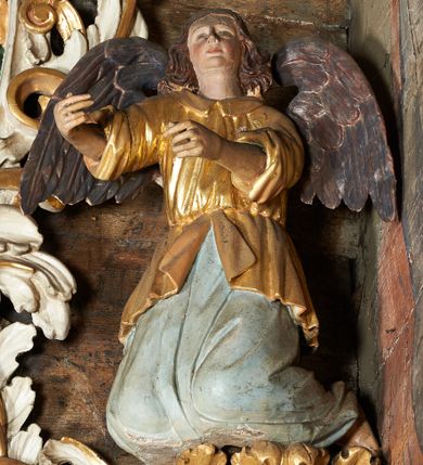 Zdjęcie nr 1: Rzeźby aniołów flankujące cokół nastawy ołtarzowej ukazane w pozycji stojącej, zwrócone przeciwstawnie, w trzech czwartych ku środkowi nastawy z wyciągniętymi doń rękami. Ubrane w długie zielone suknie, z rozcięciem odsłaniającym jedną nogę powyżej kolana oraz złoconą tunikę z podwiniętymi rękawami do łokci i kołnierzykiem. Anioły mają po jednym skrzydle, usytuowanym od strony zewnętrznej. Modelunek postaci jest sztywny. Anioły osadzone po bokach zwieńczenia ukazane są w pozycji klęczącej, przeciwstawnie do siebie, zwrócone w trzech czwartych ku środkowi, z wyciągniętymi doń rękami. Ubrane w długie jasnozielone suknie oraz złocone tuniki z podwiniętymi rękawami do łokci i kołnierzykiem. Anioły mają dwa skrzydła, szeroko rozłożone. Anioł u szczytu nastawy jest ukazany w pozycji stojącej, frontalnie, w delikatnym kontrapoście, z wyciągniętymi do przodu rękami, z głową zwróconą w prawo, z rozłożonymi szeroko skrzydłami. Ubrany analogicznie jak anioły flankujące zwieńczenie. 
Twarze wszystkich aniołów owalne, z silnie wysuniętym podbródkiem, zarumienionymi policzkami i małymi oczami. Włosy długie, ciemnobrązowe, kędzierzawe, z przedziałkiem pośrodku, opadające na plecy. Polichromia w odsłoniętych partiach ciała naturalistyczna, skrzydła srebrzone. 
