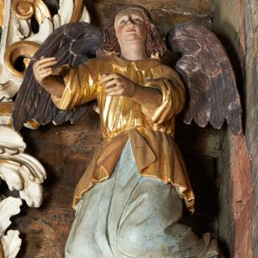 Zdjęcie nr 1: Rzeźby aniołów flankujące cokół nastawy ołtarzowej ukazane w pozycji stojącej, zwrócone przeciwstawnie, w trzech czwartych ku środkowi nastawy z wyciągniętymi doń rękami. Ubrane w długie zielone suknie, z rozcięciem odsłaniającym jedną nogę powyżej kolana oraz złoconą tunikę z podwiniętymi rękawami do łokci i kołnierzykiem. Anioły mają po jednym skrzydle, usytuowanym od strony zewnętrznej. Modelunek postaci jest sztywny. Anioły osadzone po bokach zwieńczenia ukazane są w pozycji klęczącej, przeciwstawnie do siebie, zwrócone w trzech czwartych ku środkowi, z wyciągniętymi doń rękami. Ubrane w długie jasnozielone suknie oraz złocone tuniki z podwiniętymi rękawami do łokci i kołnierzykiem. Anioły mają dwa skrzydła, szeroko rozłożone. Anioł u szczytu nastawy jest ukazany w pozycji stojącej, frontalnie, w delikatnym kontrapoście, z wyciągniętymi do przodu rękami, z głową zwróconą w prawo, z rozłożonymi szeroko skrzydłami. Ubrany analogicznie jak anioły flankujące zwieńczenie. 
Twarze wszystkich aniołów owalne, z silnie wysuniętym podbródkiem, zarumienionymi policzkami i małymi oczami. Włosy długie, ciemnobrązowe, kędzierzawe, z przedziałkiem pośrodku, opadające na plecy. Polichromia w odsłoniętych partiach ciała naturalistyczna, skrzydła srebrzone. 
