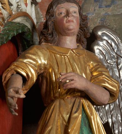 Zdjęcie nr 7: Rzeźby aniołów flankujące cokół nastawy ołtarzowej ukazane w pozycji stojącej, zwrócone przeciwstawnie, w trzech czwartych ku środkowi nastawy z wyciągniętymi doń rękami. Ubrane w długie zielone suknie, z rozcięciem odsłaniającym jedną nogę powyżej kolana oraz złoconą tunikę z podwiniętymi rękawami do łokci i kołnierzykiem. Anioły mają po jednym skrzydle, usytuowanym od strony zewnętrznej. Modelunek postaci jest sztywny. Anioły osadzone po bokach zwieńczenia ukazane są w pozycji klęczącej, przeciwstawnie do siebie, zwrócone w trzech czwartych ku środkowi, z wyciągniętymi doń rękami. Ubrane w długie jasnozielone suknie oraz złocone tuniki z podwiniętymi rękawami do łokci i kołnierzykiem. Anioły mają dwa skrzydła, szeroko rozłożone. Anioł u szczytu nastawy jest ukazany w pozycji stojącej, frontalnie, w delikatnym kontrapoście, z wyciągniętymi do przodu rękami, z głową zwróconą w prawo, z rozłożonymi szeroko skrzydłami. Ubrany analogicznie jak anioły flankujące zwieńczenie. 
Twarze wszystkich aniołów owalne, z silnie wysuniętym podbródkiem, zarumienionymi policzkami i małymi oczami. Włosy długie, ciemnobrązowe, kędzierzawe, z przedziałkiem pośrodku, opadające na plecy. Polichromia w odsłoniętych partiach ciała naturalistyczna, skrzydła srebrzone. 
