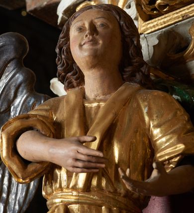 Zdjęcie nr 6: Rzeźby aniołów flankujące cokół nastawy ołtarzowej ukazane w pozycji stojącej, zwrócone przeciwstawnie, w trzech czwartych ku środkowi nastawy z wyciągniętymi doń rękami. Ubrane w długie zielone suknie, z rozcięciem odsłaniającym jedną nogę powyżej kolana oraz złoconą tunikę z podwiniętymi rękawami do łokci i kołnierzykiem. Anioły mają po jednym skrzydle, usytuowanym od strony zewnętrznej. Modelunek postaci jest sztywny. Anioły osadzone po bokach zwieńczenia ukazane są w pozycji klęczącej, przeciwstawnie do siebie, zwrócone w trzech czwartych ku środkowi, z wyciągniętymi doń rękami. Ubrane w długie jasnozielone suknie oraz złocone tuniki z podwiniętymi rękawami do łokci i kołnierzykiem. Anioły mają dwa skrzydła, szeroko rozłożone. Anioł u szczytu nastawy jest ukazany w pozycji stojącej, frontalnie, w delikatnym kontrapoście, z wyciągniętymi do przodu rękami, z głową zwróconą w prawo, z rozłożonymi szeroko skrzydłami. Ubrany analogicznie jak anioły flankujące zwieńczenie. 
Twarze wszystkich aniołów owalne, z silnie wysuniętym podbródkiem, zarumienionymi policzkami i małymi oczami. Włosy długie, ciemnobrązowe, kędzierzawe, z przedziałkiem pośrodku, opadające na plecy. Polichromia w odsłoniętych partiach ciała naturalistyczna, skrzydła srebrzone. 
