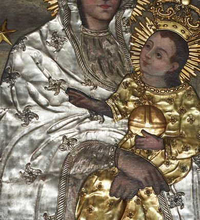 Zdjęcie nr 1: Obraz w kształcie stojącego prostokąta, zamkniętego łukiem wklęsło-wypukłym z uskokami, z wizerunkiem Matki Boskiej Śnieżnej. Maria przedstawiona frontalnie, w półpostaci, z Dzieciątkiem Jezus na lewym ręku, ze skrzyżowanymi dłońmi. Twarz podłużna z długim i wąskim nosem, drobnymi ustami, dużymi oczami obwiedzionymi regularnymi łukami brwiowymi, ze wzrokiem skierowanym wprost na widza. Dzieciątko zwrócone trzy czwarte w prawo, w pozycji siedzącej, z jabłkiem królewskim w lewej dłoni, prawą czyniące gest błogosławieństwa, na stopach ma założone sandałki. Twarz okrągła o wyrazistych rysach, duże i brązowe oczy, zarumienione policzki, włosy średniej długości opadające na plecy, wzrok skierowany na Matkę. Tło w odcieniach szarości, w górnej części pochmurne niebo. Nad głową Marii gołębica Ducha Świętego z kwiatem róży w dziobie. W górnych narożach dwie przeciwstawnie ustawione, uskrzydlone główki aniołków. Na postacie Marii i Dzieciątka nałożone metalowe sukienki, promieniste nimby, korony w typie zamkniętym i wieniec z 12 gwiazd wokół głów.
