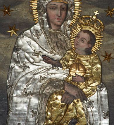 Zdjęcie nr 4: Obraz w kształcie stojącego prostokąta, zamkniętego łukiem wklęsło-wypukłym z uskokami, z wizerunkiem Matki Boskiej Śnieżnej. Maria przedstawiona frontalnie, w półpostaci, z Dzieciątkiem Jezus na lewym ręku, ze skrzyżowanymi dłońmi. Twarz podłużna z długim i wąskim nosem, drobnymi ustami, dużymi oczami obwiedzionymi regularnymi łukami brwiowymi, ze wzrokiem skierowanym wprost na widza. Dzieciątko zwrócone trzy czwarte w prawo, w pozycji siedzącej, z jabłkiem królewskim w lewej dłoni, prawą czyniące gest błogosławieństwa, na stopach ma założone sandałki. Twarz okrągła o wyrazistych rysach, duże i brązowe oczy, zarumienione policzki, włosy średniej długości opadające na plecy, wzrok skierowany na Matkę. Tło w odcieniach szarości, w górnej części pochmurne niebo. Nad głową Marii gołębica Ducha Świętego z kwiatem róży w dziobie. W górnych narożach dwie przeciwstawnie ustawione, uskrzydlone główki aniołków. Na postacie Marii i Dzieciątka nałożone metalowe sukienki, promieniste nimby, korony w typie zamkniętym i wieniec z 12 gwiazd wokół głów.
