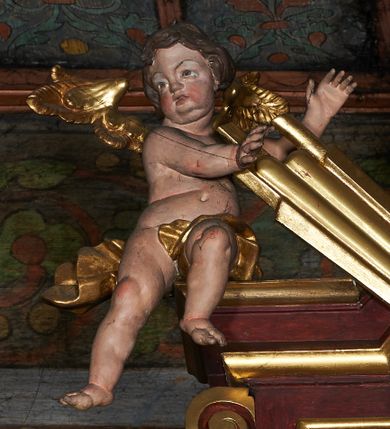 Zdjęcie nr 1: Rzeźby aniołków na segmentowych przyczółkach ukazane w pozycji klęczącej, natomiast rzeźby w partii zwieńczenia w pozycji siedzącej. Wszystkie figury zwrócone ku sobie, przeciwstawnie, z głowami uniesionym do góry, z rękami wyrażającymi teatralne gesty, w pasie przewiązane złotą chustą; modelunek ciała subtelny. Twarze okrągłe, z pucułowatymi policzkami i otwartymi ustami. Włosy średniej długości, ciemnobrązowe, zasłaniające uszy. Polichromia w odsłoniętych partiach ciała naturalistyczna, skrzydła i chusty złocone. 
