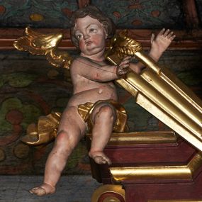 Zdjęcie nr 1: Rzeźby aniołków na segmentowych przyczółkach ukazane w pozycji klęczącej, natomiast rzeźby w partii zwieńczenia w pozycji siedzącej. Wszystkie figury zwrócone ku sobie, przeciwstawnie, z głowami uniesionym do góry, z rękami wyrażającymi teatralne gesty, w pasie przewiązane złotą chustą; modelunek ciała subtelny. Twarze okrągłe, z pucułowatymi policzkami i otwartymi ustami. Włosy średniej długości, ciemnobrązowe, zasłaniające uszy. Polichromia w odsłoniętych partiach ciała naturalistyczna, skrzydła i chusty złocone. 
