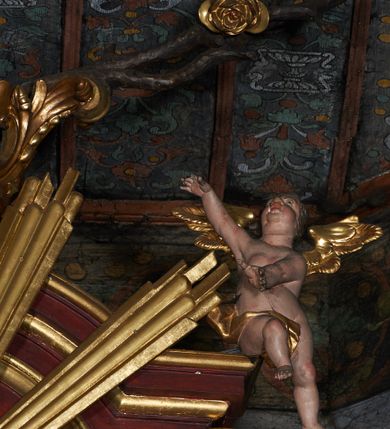 Zdjęcie nr 4: Rzeźby aniołków na segmentowych przyczółkach ukazane w pozycji klęczącej, natomiast rzeźby w partii zwieńczenia w pozycji siedzącej. Wszystkie figury zwrócone ku sobie, przeciwstawnie, z głowami uniesionym do góry, z rękami wyrażającymi teatralne gesty, w pasie przewiązane złotą chustą; modelunek ciała subtelny. Twarze okrągłe, z pucułowatymi policzkami i otwartymi ustami. Włosy średniej długości, ciemnobrązowe, zasłaniające uszy. Polichromia w odsłoniętych partiach ciała naturalistyczna, skrzydła i chusty złocone. 
