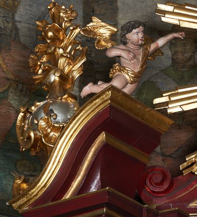Zdjęcie nr 5: Rzeźby aniołków na segmentowych przyczółkach ukazane w pozycji klęczącej, natomiast rzeźby w partii zwieńczenia w pozycji siedzącej. Wszystkie figury zwrócone ku sobie, przeciwstawnie, z głowami uniesionym do góry, z rękami wyrażającymi teatralne gesty, w pasie przewiązane złotą chustą; modelunek ciała subtelny. Twarze okrągłe, z pucułowatymi policzkami i otwartymi ustami. Włosy średniej długości, ciemnobrązowe, zasłaniające uszy. Polichromia w odsłoniętych partiach ciała naturalistyczna, skrzydła i chusty złocone. 
