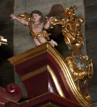 Zdjęcie nr 6: Rzeźby aniołków na segmentowych przyczółkach ukazane w pozycji klęczącej, natomiast rzeźby w partii zwieńczenia w pozycji siedzącej. Wszystkie figury zwrócone ku sobie, przeciwstawnie, z głowami uniesionym do góry, z rękami wyrażającymi teatralne gesty, w pasie przewiązane złotą chustą; modelunek ciała subtelny. Twarze okrągłe, z pucułowatymi policzkami i otwartymi ustami. Włosy średniej długości, ciemnobrązowe, zasłaniające uszy. Polichromia w odsłoniętych partiach ciała naturalistyczna, skrzydła i chusty złocone. 
