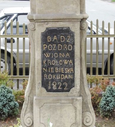 Zdjęcie nr 2: Kapliczka wolnostojąca z postacią Matki Boskiej Różańcowej na wysokim cokole. Podstawa podzielona na trzy strefy – dolna na uskokowym cokole z płyciną w kształcie leżącego prostokąta o wklęsłych narożach, wyższa z tablicą z kutym, złoconym napisem „BĄDŹ / POZDRO/WIONA / KRÓLOWA / NIEBIESKA / ROKU PAN(SKIEGO) / 1822”, najwyższa gładka. Całość ujęta spływami wolutowymi i zwieńczona belkowaniem przechodzącym na osi w łuk nadwieszony z wpisaną palmetą. Na cokole figura Matki Boskiej ukazanej całopostaciowo, frontalnie z Dzieciątkiem na lewym ramieniu, prawą ręką trzyma ujęty przez nie różaniec. Obie postaci mają na głowie zamknięte korony. Maria jest ubrana w białą suknię i niebieski płaszcz, Dzieciątko w czerwoną sukienkę. Postacie przykrywa metalowy daszek wsparty na czterech prętach. 