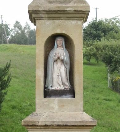 Zdjęcie nr 2: Kapliczka wolnostojąca z figurą św. Floriana na wysokim, dwustrefowym cokole. Dolna część prostopadłościenna, na profilowanej podstawie, z płytką niszą w kształcie stojącego prostokąta zamkniętego łukiem pełnym na każdej ścianie. Na froncie napis „TA PAMIATKA / Małżonków Marcina i Telke / Zapałowiczów w roku 1882 / Fundatorowie proszą / przechodnich o westchnienie / za dusze Zmarłych”. Na bocznych ścianach płaskorzeźbione postacie świętych ukazanych w całej postaci, frontalnie. Z jednej strony wizerunek  świętej męczennicy z płonącą budowlą z lewej strony i psem/smokiem z prawej (św. Małgorzata?). Święta trzyma w prawej ręce palmę męczeństwa, lewą przyciska do piersi. Ma łagodną, owalną twarz, włosy z przedziałkiem, zaczesane do tyłu. Jest ubrana w czerwoną, przepasaną suknię i czerwony płaszcz.  Na innej płaszczyźnie postać św. Andrzeja przedstawionego jako brodaty mężczyzna w niebieskiej tunice i czerwonym płaszczu. Za jego plecami widać końce ramion krzyża w kształcie litery „X”. Na kolejnej św. Jan Nepomucen w sutannie, komży i płaszczu, w birecie. Z krucyfiksem i palmą męczeństwa w rękach. Na kolejnej ściance św. Marcin na koniu, w stroju żołnierza, trzymanym w prawej ręce mieczem odcina część płaszcza, który chwyta siedząca na ziemi postać w czerwonych spodniach i zielonej koszuli. W górnej strefie cokołu, w ścianie frontowej nisza z figurą Matki Boskiej w całej postaci, frontalnie, z rękami złożonymi do modlitwy i różańcem. Maria ma na sobie białą suknię i niebieski płaszcz zarzucony na głowę; na bosych stopach kwiaty róż Na cokole postać św. Floriana w całej postaci, frontalnie, z cebrzykiem z którego leje się woda na płonący dom w prawej i włócznią w lewej ręce. Ma owalną twarz, wąsy i brodę. Ubrany jest w strój rzymskiego legionisty – jasną tunikę, czerwony płaszcz, wysokie buty i hełm.