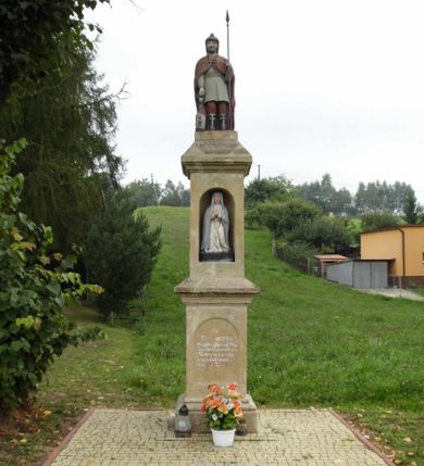 Zdjęcie nr 1: Kapliczka wolnostojąca z figurą św. Floriana na wysokim, dwustrefowym cokole. Dolna część prostopadłościenna, na profilowanej podstawie, z płytką niszą w kształcie stojącego prostokąta zamkniętego łukiem pełnym na każdej ścianie. Na froncie napis „TA PAMIATKA / Małżonków Marcina i Telke / Zapałowiczów w roku 1882 / Fundatorowie proszą / przechodnich o westchnienie / za dusze Zmarłych”. Na bocznych ścianach płaskorzeźbione postacie świętych ukazanych w całej postaci, frontalnie. Z jednej strony wizerunek  świętej męczennicy z płonącą budowlą z lewej strony i psem/smokiem z prawej (św. Małgorzata?). Święta trzyma w prawej ręce palmę męczeństwa, lewą przyciska do piersi. Ma łagodną, owalną twarz, włosy z przedziałkiem, zaczesane do tyłu. Jest ubrana w czerwoną, przepasaną suknię i czerwony płaszcz.  Na innej płaszczyźnie postać św. Andrzeja przedstawionego jako brodaty mężczyzna w niebieskiej tunice i czerwonym płaszczu. Za jego plecami widać końce ramion krzyża w kształcie litery „X”. Na kolejnej św. Jan Nepomucen w sutannie, komży i płaszczu, w birecie. Z krucyfiksem i palmą męczeństwa w rękach. Na kolejnej ściance św. Marcin na koniu, w stroju żołnierza, trzymanym w prawej ręce mieczem odcina część płaszcza, który chwyta siedząca na ziemi postać w czerwonych spodniach i zielonej koszuli. W górnej strefie cokołu, w ścianie frontowej nisza z figurą Matki Boskiej w całej postaci, frontalnie, z rękami złożonymi do modlitwy i różańcem. Maria ma na sobie białą suknię i niebieski płaszcz zarzucony na głowę; na bosych stopach kwiaty róż Na cokole postać św. Floriana w całej postaci, frontalnie, z cebrzykiem z którego leje się woda na płonący dom w prawej i włócznią w lewej ręce. Ma owalną twarz, wąsy i brodę. Ubrany jest w strój rzymskiego legionisty – jasną tunikę, czerwony płaszcz, wysokie buty i hełm.