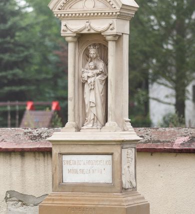 Zdjęcie nr 2: Kapliczka wolnostojąca na wysokim, dwustrefowym cokole. Dolna część szersza, dzielona żłobieniem, z napisem w górnej części „FUNDAROR / SZYMON KOSCIELNY / 1888 r(ok)”. Powyżej węższa, prostopadłościenna część z wyrytą na froncie na kamiennej, wprawionej tablicy inskrypcją „ŚWIĘTA BOŻA RODZICIELKO / MÓDL SIĘ ZA NAMI”. Na prawej ściance bocznej płaskorzeźba z półpostacią św. Szymona Apostoła. Święty zwrócony lekko w prawo, z głową skierowaną w trzech czwartych w tę samą stronę. Ma pociągłą twarz z głęboko osadzonymi oczami, wąskim nosem, niewielkimi ustami przesłoniętymi częściowo wąsami. Twarz okalają zwinięte w pukle włosy i dzieląca się na dwie części broda. Święty lewą ręką przytrzymuje książkę, prawą piłę. Jest ubrany w tunikę z długimi rękawami. Po przeciwnej stronie cokołu podobizna św. Wojciecha ukazanego w podobny sposób. Święty jest ubrany w strój biskupa – komżę, kapę, paliusz i infułę. W prawej ręce trzyma krzyż patriarszy oraz wiosło, lewą przytrzymuje zamkniętą księgę.  Na postumencie ustawiono figurę Marii z Dzieciątkiem w niszy zwieńczonej przyczółkiem wspartym na dwóch kolumnach wychodzących przed jej lico. Maria została przedstawiona całopostaciowo, frontalnie, podtrzymując obiema rękami Dzieciątko, które rozpościera ramiona. Maria ma delikatną, owalną twarz o półprzymkniętych oczach, wąskim nosie i cofniętej brodzie. Długie włosy spływają jej na plecy i ramiona. Jest ubrana w długą suknię i płaszcz z ornamentalna bordiurą. Na głowie ma otwartą koronę. Dzieciątko jest ubrane w tunikę do kolan, ma owalną twarz z głęboko osadzonymi oczami i małym nosem i ustami; włosy opadają puklami na czoło. Niszę wypełnia muszla palmetowa, na gzymsie belkowania rzeźbiona wstęga przepleciona przez uchwyt, gzyms dekorowany kostkowaniem. Półokrągły tympanon wypełniają trzy płyciny – środkowa okrągła, boczne trójkątne o wklęsło-wypukłych krawędziach, powyżej kostkowanie. Na tympanonie, na cokole krucyfiks z wypukle rzeźbioną postacią Chrystusa i titulusem z napisem „INRI”. 