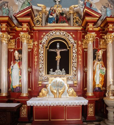 Zdjęcie nr 12: Ołtarz architektoniczny, trójosiowy, jednokondygnacyjny ze zwieńczeniem, na cokole. Mensa prostopadłościenna, drewniana, z portatylem. Antepedium na profilowanej podstawie podzielone na trzy bliskie kwadratom płyciny. Metalowe tabernakulum w formie prostopadłościanu ujętego wolutami z liści. Na ścianie frontowej drzwiczki dekorowane hostią z literami IHS ujętą od dołu rozłożonymi dłońmi.  Osie nastawy wydzielone dwiema parami pilastrów i stojącymi przed licem ołtarza kolumnami z kapitelami kompozytowymi. Wewnętrzna para kolumn dźwiga imposty z przerwanym przyczółkiem, na którym siedzi para klęczących aniołów, zaś zewnętrzna para dźwiga imposty z aniołami półleżącymi. Pole środkowe tworzy szeroka, wypełniona ażurowym akantem i rozetami rama w formie stojącego prostokąta zamkniętego łukiem nadwieszonym, ujęta lizenami, podtrzymywana przez konsole, z obrazem św. Wawrzyńca wymiennie z obrazem św. Józefa oraz rzeźbionym krucyfiksem. W osiach bocznych figury św. Stanisława po lewej i św. Wojciecha po prawej stronie, na tle arkad z ażurową kratką. Zwieńczenie tworzy grupa rzeźbiarska z przedstawieniem Trójcy Świętej. Bóg Ojciec i Chrystus siedzą zwróceni do siebie, na tle ulistionych wolut, które podtrzymują gołębicę Ducha Świetego, nad którą unosi się Oko Opatrzności. Grupa jest flankowana przez jońskie kolumny ze zwróconymi na zewnątrz wolutami, ujęta fragmentami wici tworzącymi spływy wolutowe. Cokoły kolumn dekorowane płycinami z akantem, rozetą i liliami heraldycznymi, zwieńczone ząbkowaniem. W przyłuczach dekoracja roślinna. Płycinę lizen zdobią festony z wstęgą regencyjną. Trzony pilastrów podzielone na dwie płyciny o wklęsłych bokach, dekorowane rozetami. Architrawy i fryzy dekorowane rozetami z akantu, gzyms z ząbkowaniem. Kolumny zwieńczenia dekorowane akantowymi spływami wolutowymi. Ołtarz polichromowany na ciemnoczerwono; ornamentyka oraz detale architektoniczne złocone i srebrzone. 