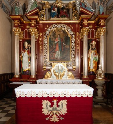Zdjęcie nr 3: Ołtarz architektoniczny, trójosiowy, jednokondygnacyjny ze zwieńczeniem, na cokole. Mensa prostopadłościenna, drewniana, z portatylem. Antepedium na profilowanej podstawie podzielone na trzy bliskie kwadratom płyciny. Metalowe tabernakulum w formie prostopadłościanu ujętego wolutami z liści. Na ścianie frontowej drzwiczki dekorowane hostią z literami IHS ujętą od dołu rozłożonymi dłońmi.  Osie nastawy wydzielone dwiema parami pilastrów i stojącymi przed licem ołtarza kolumnami z kapitelami kompozytowymi. Wewnętrzna para kolumn dźwiga imposty z przerwanym przyczółkiem, na którym siedzi para klęczących aniołów, zaś zewnętrzna para dźwiga imposty z aniołami półleżącymi. Pole środkowe tworzy szeroka, wypełniona ażurowym akantem i rozetami rama w formie stojącego prostokąta zamkniętego łukiem nadwieszonym, ujęta lizenami, podtrzymywana przez konsole, z obrazem św. Wawrzyńca wymiennie z obrazem św. Józefa oraz rzeźbionym krucyfiksem. W osiach bocznych figury św. Stanisława po lewej i św. Wojciecha po prawej stronie, na tle arkad z ażurową kratką. Zwieńczenie tworzy grupa rzeźbiarska z przedstawieniem Trójcy Świętej. Bóg Ojciec i Chrystus siedzą zwróceni do siebie, na tle ulistionych wolut, które podtrzymują gołębicę Ducha Świetego, nad którą unosi się Oko Opatrzności. Grupa jest flankowana przez jońskie kolumny ze zwróconymi na zewnątrz wolutami, ujęta fragmentami wici tworzącymi spływy wolutowe. Cokoły kolumn dekorowane płycinami z akantem, rozetą i liliami heraldycznymi, zwieńczone ząbkowaniem. W przyłuczach dekoracja roślinna. Płycinę lizen zdobią festony z wstęgą regencyjną. Trzony pilastrów podzielone na dwie płyciny o wklęsłych bokach, dekorowane rozetami. Architrawy i fryzy dekorowane rozetami z akantu, gzyms z ząbkowaniem. Kolumny zwieńczenia dekorowane akantowymi spływami wolutowymi. Ołtarz polichromowany na ciemnoczerwono; ornamentyka oraz detale architektoniczne złocone i srebrzone. 
