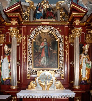 Zdjęcie nr 2: Ołtarz architektoniczny, trójosiowy, jednokondygnacyjny ze zwieńczeniem, na cokole. Mensa prostopadłościenna, drewniana, z portatylem. Antepedium na profilowanej podstawie podzielone na trzy bliskie kwadratom płyciny. Metalowe tabernakulum w formie prostopadłościanu ujętego wolutami z liści. Na ścianie frontowej drzwiczki dekorowane hostią z literami IHS ujętą od dołu rozłożonymi dłońmi.  Osie nastawy wydzielone dwiema parami pilastrów i stojącymi przed licem ołtarza kolumnami z kapitelami kompozytowymi. Wewnętrzna para kolumn dźwiga imposty z przerwanym przyczółkiem, na którym siedzi para klęczących aniołów, zaś zewnętrzna para dźwiga imposty z aniołami półleżącymi. Pole środkowe tworzy szeroka, wypełniona ażurowym akantem i rozetami rama w formie stojącego prostokąta zamkniętego łukiem nadwieszonym, ujęta lizenami, podtrzymywana przez konsole, z obrazem św. Wawrzyńca wymiennie z obrazem św. Józefa oraz rzeźbionym krucyfiksem. W osiach bocznych figury św. Stanisława po lewej i św. Wojciecha po prawej stronie, na tle arkad z ażurową kratką. Zwieńczenie tworzy grupa rzeźbiarska z przedstawieniem Trójcy Świętej. Bóg Ojciec i Chrystus siedzą zwróceni do siebie, na tle ulistionych wolut, które podtrzymują gołębicę Ducha Świetego, nad którą unosi się Oko Opatrzności. Grupa jest flankowana przez jońskie kolumny ze zwróconymi na zewnątrz wolutami, ujęta fragmentami wici tworzącymi spływy wolutowe. Cokoły kolumn dekorowane płycinami z akantem, rozetą i liliami heraldycznymi, zwieńczone ząbkowaniem. W przyłuczach dekoracja roślinna. Płycinę lizen zdobią festony z wstęgą regencyjną. Trzony pilastrów podzielone na dwie płyciny o wklęsłych bokach, dekorowane rozetami. Architrawy i fryzy dekorowane rozetami z akantu, gzyms z ząbkowaniem. Kolumny zwieńczenia dekorowane akantowymi spływami wolutowymi. Ołtarz polichromowany na ciemnoczerwono; ornamentyka oraz detale architektoniczne złocone i srebrzone. 
