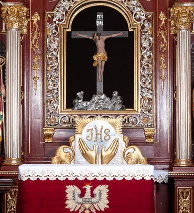 Zdjęcie nr 16: Ołtarz architektoniczny, trójosiowy, jednokondygnacyjny ze zwieńczeniem, na cokole. Mensa prostopadłościenna, drewniana, z portatylem. Antepedium na profilowanej podstawie podzielone na trzy bliskie kwadratom płyciny. Metalowe tabernakulum w formie prostopadłościanu ujętego wolutami z liści. Na ścianie frontowej drzwiczki dekorowane hostią z literami IHS ujętą od dołu rozłożonymi dłońmi.  Osie nastawy wydzielone dwiema parami pilastrów i stojącymi przed licem ołtarza kolumnami z kapitelami kompozytowymi. Wewnętrzna para kolumn dźwiga imposty z przerwanym przyczółkiem, na którym siedzi para klęczących aniołów, zaś zewnętrzna para dźwiga imposty z aniołami półleżącymi. Pole środkowe tworzy szeroka, wypełniona ażurowym akantem i rozetami rama w formie stojącego prostokąta zamkniętego łukiem nadwieszonym, ujęta lizenami, podtrzymywana przez konsole, z obrazem św. Wawrzyńca wymiennie z obrazem św. Józefa oraz rzeźbionym krucyfiksem. W osiach bocznych figury św. Stanisława po lewej i św. Wojciecha po prawej stronie, na tle arkad z ażurową kratką. Zwieńczenie tworzy grupa rzeźbiarska z przedstawieniem Trójcy Świętej. Bóg Ojciec i Chrystus siedzą zwróceni do siebie, na tle ulistionych wolut, które podtrzymują gołębicę Ducha Świetego, nad którą unosi się Oko Opatrzności. Grupa jest flankowana przez jońskie kolumny ze zwróconymi na zewnątrz wolutami, ujęta fragmentami wici tworzącymi spływy wolutowe. Cokoły kolumn dekorowane płycinami z akantem, rozetą i liliami heraldycznymi, zwieńczone ząbkowaniem. W przyłuczach dekoracja roślinna. Płycinę lizen zdobią festony z wstęgą regencyjną. Trzony pilastrów podzielone na dwie płyciny o wklęsłych bokach, dekorowane rozetami. Architrawy i fryzy dekorowane rozetami z akantu, gzyms z ząbkowaniem. Kolumny zwieńczenia dekorowane akantowymi spływami wolutowymi. Ołtarz polichromowany na ciemnoczerwono; ornamentyka oraz detale architektoniczne złocone i srebrzone. 