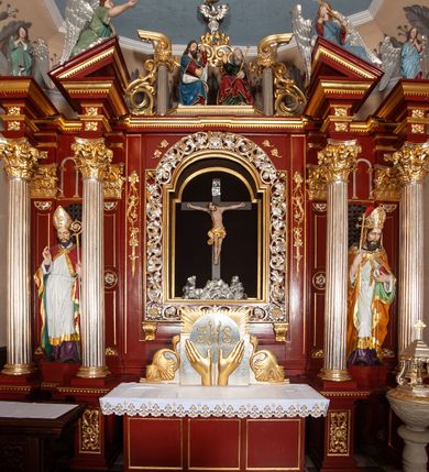 Zdjęcie nr 13: Ołtarz architektoniczny, trójosiowy, jednokondygnacyjny ze zwieńczeniem, na cokole. Mensa prostopadłościenna, drewniana, z portatylem. Antepedium na profilowanej podstawie podzielone na trzy bliskie kwadratom płyciny. Metalowe tabernakulum w formie prostopadłościanu ujętego wolutami z liści. Na ścianie frontowej drzwiczki dekorowane hostią z literami IHS ujętą od dołu rozłożonymi dłońmi.  Osie nastawy wydzielone dwiema parami pilastrów i stojącymi przed licem ołtarza kolumnami z kapitelami kompozytowymi. Wewnętrzna para kolumn dźwiga imposty z przerwanym przyczółkiem, na którym siedzi para klęczących aniołów, zaś zewnętrzna para dźwiga imposty z aniołami półleżącymi. Pole środkowe tworzy szeroka, wypełniona ażurowym akantem i rozetami rama w formie stojącego prostokąta zamkniętego łukiem nadwieszonym, ujęta lizenami, podtrzymywana przez konsole, z obrazem św. Wawrzyńca wymiennie z obrazem św. Józefa oraz rzeźbionym krucyfiksem. W osiach bocznych figury św. Stanisława po lewej i św. Wojciecha po prawej stronie, na tle arkad z ażurową kratką. Zwieńczenie tworzy grupa rzeźbiarska z przedstawieniem Trójcy Świętej. Bóg Ojciec i Chrystus siedzą zwróceni do siebie, na tle ulistionych wolut, które podtrzymują gołębicę Ducha Świetego, nad którą unosi się Oko Opatrzności. Grupa jest flankowana przez jońskie kolumny ze zwróconymi na zewnątrz wolutami, ujęta fragmentami wici tworzącymi spływy wolutowe. Cokoły kolumn dekorowane płycinami z akantem, rozetą i liliami heraldycznymi, zwieńczone ząbkowaniem. W przyłuczach dekoracja roślinna. Płycinę lizen zdobią festony z wstęgą regencyjną. Trzony pilastrów podzielone na dwie płyciny o wklęsłych bokach, dekorowane rozetami. Architrawy i fryzy dekorowane rozetami z akantu, gzyms z ząbkowaniem. Kolumny zwieńczenia dekorowane akantowymi spływami wolutowymi. Ołtarz polichromowany na ciemnoczerwono; ornamentyka oraz detale architektoniczne złocone i srebrzone. 