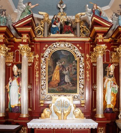 Zdjęcie nr 20: Ołtarz architektoniczny, trójosiowy, jednokondygnacyjny ze zwieńczeniem, na cokole. Mensa prostopadłościenna, drewniana, z portatylem. Antepedium na profilowanej podstawie podzielone na trzy bliskie kwadratom płyciny. Metalowe tabernakulum w formie prostopadłościanu ujętego wolutami z liści. Na ścianie frontowej drzwiczki dekorowane hostią z literami IHS ujętą od dołu rozłożonymi dłońmi.  Osie nastawy wydzielone dwiema parami pilastrów i stojącymi przed licem ołtarza kolumnami z kapitelami kompozytowymi. Wewnętrzna para kolumn dźwiga imposty z przerwanym przyczółkiem, na którym siedzi para klęczących aniołów, zaś zewnętrzna para dźwiga imposty z aniołami półleżącymi. Pole środkowe tworzy szeroka, wypełniona ażurowym akantem i rozetami rama w formie stojącego prostokąta zamkniętego łukiem nadwieszonym, ujęta lizenami, podtrzymywana przez konsole, z obrazem św. Wawrzyńca wymiennie z obrazem św. Józefa oraz rzeźbionym krucyfiksem. W osiach bocznych figury św. Stanisława po lewej i św. Wojciecha po prawej stronie, na tle arkad z ażurową kratką. Zwieńczenie tworzy grupa rzeźbiarska z przedstawieniem Trójcy Świętej. Bóg Ojciec i Chrystus siedzą zwróceni do siebie, na tle ulistionych wolut, które podtrzymują gołębicę Ducha Świetego, nad którą unosi się Oko Opatrzności. Grupa jest flankowana przez jońskie kolumny ze zwróconymi na zewnątrz wolutami, ujęta fragmentami wici tworzącymi spływy wolutowe. Cokoły kolumn dekorowane płycinami z akantem, rozetą i liliami heraldycznymi, zwieńczone ząbkowaniem. W przyłuczach dekoracja roślinna. Płycinę lizen zdobią festony z wstęgą regencyjną. Trzony pilastrów podzielone na dwie płyciny o wklęsłych bokach, dekorowane rozetami. Architrawy i fryzy dekorowane rozetami z akantu, gzyms z ząbkowaniem. Kolumny zwieńczenia dekorowane akantowymi spływami wolutowymi. Ołtarz polichromowany na ciemnoczerwono; ornamentyka oraz detale architektoniczne złocone i srebrzone. 