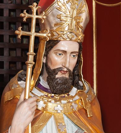 Zdjęcie nr 1: Figura drążona, na cokole. Święty Wojciech został ukazany w pozycji stojącej z uniesionymi rękami, w prawej trzyma podwójny krzyż misyjny, w lewej zaś, przy piersi, oprawioną w czerwień księgę z zaznaczonym palcem fragmentem. Twarz ma pociągłą, brwi wąskie, oczy szeroko otwarte, nos prosty, wąski i długą, sięgającą dekoltu brodę. Postać jest ubrana w fioletową albę, białą komżę ze złoconą bordiurą i ściśle przylegającą do ciała pomarańczową kapę, podbitą zielenią. Na nią ma załozony złoty paliusz, na głowie infułę, na nogach ciemnobrązowe buty. Draperia delikatnie fałdowana, odzwierciedla układ ciała. W partiach ciała polichromia naturalistyczna.