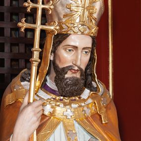 Zdjęcie nr 1: Figura drążona, na cokole. Święty Wojciech został ukazany w pozycji stojącej z uniesionymi rękami, w prawej trzyma podwójny krzyż misyjny, w lewej zaś, przy piersi, oprawioną w czerwień księgę z zaznaczonym palcem fragmentem. Twarz ma pociągłą, brwi wąskie, oczy szeroko otwarte, nos prosty, wąski i długą, sięgającą dekoltu brodę. Postać jest ubrana w fioletową albę, białą komżę ze złoconą bordiurą i ściśle przylegającą do ciała pomarańczową kapę, podbitą zielenią. Na nią ma załozony złoty paliusz, na głowie infułę, na nogach ciemnobrązowe buty. Draperia delikatnie fałdowana, odzwierciedla układ ciała. W partiach ciała polichromia naturalistyczna.