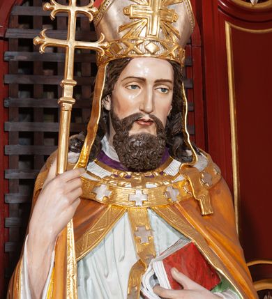 Zdjęcie nr 4: Figura drążona, na cokole. Święty Wojciech został ukazany w pozycji stojącej z uniesionymi rękami, w prawej trzyma podwójny krzyż misyjny, w lewej zaś, przy piersi, oprawioną w czerwień księgę z zaznaczonym palcem fragmentem. Twarz ma pociągłą, brwi wąskie, oczy szeroko otwarte, nos prosty, wąski i długą, sięgającą dekoltu brodę. Postać jest ubrana w fioletową albę, białą komżę ze złoconą bordiurą i ściśle przylegającą do ciała pomarańczową kapę, podbitą zielenią. Na nią ma załozony złoty paliusz, na głowie infułę, na nogach ciemnobrązowe buty. Draperia delikatnie fałdowana, odzwierciedla układ ciała. W partiach ciała polichromia naturalistyczna.