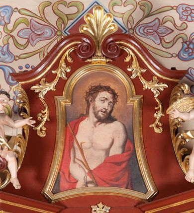 Zdjęcie nr 12: Ołtarz przyścienny, architektoniczny, jednoosiowy, jednokondygnacjowy ze zwieńczeniem. Mensa prostopadłościenna, antepedium ujęte ramą, z monogramem maryjnym na środku. Retabulum ustawione na wysokim cokole, przed którym stoi prostopadłościenna mensa. Główne pole retabulum flankują kolumny ustawione na tle filarów. Podpory mają głowice kompozytowe i są posadowione na ukośnie ustawionych, rozsuniętych postumentach dekorowanych płycinami. Dźwigają  odcinki belkowania także ustawione skośnie. W prostokątnym polu głównym nastawy ołtarzowej obraz z przedstawieniem Matki Boskiej z Dzieciątkiem, wymiennie z płótnem przedstawiającym Najświętsze Serce Jezusa i św. Stanisława Kostkę w prostokątnej ramie zamkniętej od góry łukiem półokrągłym. Na fryzie belkowania umieszczono rozety. Na zewnętrznych odcinkach belkowania, na osi kolumn, skierowane do wewnątrz woluty; na nich siedzą anioły prezentujące obraz zwieńczenia. Zwieńczenie w formie wklęsło-wypukłej, wypełnione wizerunkiem Ecce Homo we wklęsło-wypukłej ramie z łukiem nadwieszonym, jest zamknięte od góry dwiema wolutami. W miejscu ich styku, na osi palmeta. Struktura ołtarza polichromowana na brązowo; kolumny i płyciny z imitacją marmoryzacji, jasnoczerwone;  profile,  detale i ornamenty złocone. 