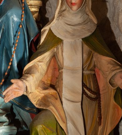 Zdjęcie nr 4: Rzeźba przedstawiająca  Matkę Boską Różańcową ze św. Dominkiem i św. Katarzyną Sieneńską. Kompozycja sceny zamyka się w trójkącie. Jej szczyt stanowi siedząca na złotym, ozdobionym ornamentami i główkami anielskimi tronie Matka Boska. Maria patrzy przed siebie i wyciągniętą prawą ręką podaje różaniec św. Dominikowi. Lewą ręką przytrzymuje Dzieciątko, które lewą ręką podaje różaniec św. Katarzynie, a prawą go podtrzymuje. Wszystkie osoby są połączone jednym sznurem różańca. Maria ma owalną, pełną twarz o jasnoniebieskich oczach, szerokim nosie i pełnych ustach. Jest ubrana w jasnoniebieskią suknię z haftowanym gorsetem i niebieski, podbity czerwienią płaszcz, który zakrywa jej lewe kolano i plecy; na głowie ma ozdobioną koronką, białą chustę, na szyi pięć sznurów czerwonych korali, na nogach sandały. Dzieciątko ma okrągłą twarz, małe oczy, nos i usta i delikatny rumieniec. Jest przewiązane białą tkaniną. Na głowach obu postaci otwarte korony. Po prawej stronie klęczy na lewym kolanie, ustawiona skosem św. Katarzyna. Prawą rękę wyciąga przed siebie, ma wąską, podłużną twarz, z niewielkimi, ciemnymi oczami, długim nosem i niewielkimi ustami; na głowie koronę cierniową. Jest ubrana w jasny habit z białym szkaplerzem, zielony, podbity jasnym kolorem płaszcz, podwikę i welon, za pas ma zatkniety różaniec. Klęczący po przeciwnej stronie kompozycji św. Dominik jest tylko częściowo widoczny. Patrzy na widza, lewą ręką przytrzymuje przekazywany przez Marię różaniec. Jest ubrany w biały habit i czarną pelerynkę, przy sznurze ma różaniec. Tło przedstawienia stanowi kamienna ściana. 