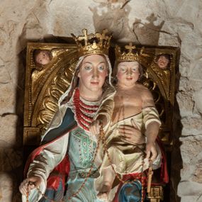 Zdjęcie nr 1: Rzeźba przedstawiająca  Matkę Boską Różańcową ze św. Dominkiem i św. Katarzyną Sieneńską. Kompozycja sceny zamyka się w trójkącie. Jej szczyt stanowi siedząca na złotym, ozdobionym ornamentami i główkami anielskimi tronie Matka Boska. Maria patrzy przed siebie i wyciągniętą prawą ręką podaje różaniec św. Dominikowi. Lewą ręką przytrzymuje Dzieciątko, które lewą ręką podaje różaniec św. Katarzynie, a prawą go podtrzymuje. Wszystkie osoby są połączone jednym sznurem różańca. Maria ma owalną, pełną twarz o jasnoniebieskich oczach, szerokim nosie i pełnych ustach. Jest ubrana w jasnoniebieskią suknię z haftowanym gorsetem i niebieski, podbity czerwienią płaszcz, który zakrywa jej lewe kolano i plecy; na głowie ma ozdobioną koronką, białą chustę, na szyi pięć sznurów czerwonych korali, na nogach sandały. Dzieciątko ma okrągłą twarz, małe oczy, nos i usta i delikatny rumieniec. Jest przewiązane białą tkaniną. Na głowach obu postaci otwarte korony. Po prawej stronie klęczy na lewym kolanie, ustawiona skosem św. Katarzyna. Prawą rękę wyciąga przed siebie, ma wąską, podłużną twarz, z niewielkimi, ciemnymi oczami, długim nosem i niewielkimi ustami; na głowie koronę cierniową. Jest ubrana w jasny habit z białym szkaplerzem, zielony, podbity jasnym kolorem płaszcz, podwikę i welon, za pas ma zatkniety różaniec. Klęczący po przeciwnej stronie kompozycji św. Dominik jest tylko częściowo widoczny. Patrzy na widza, lewą ręką przytrzymuje przekazywany przez Marię różaniec. Jest ubrany w biały habit i czarną pelerynkę, przy sznurze ma różaniec. Tło przedstawienia stanowi kamienna ściana. 
