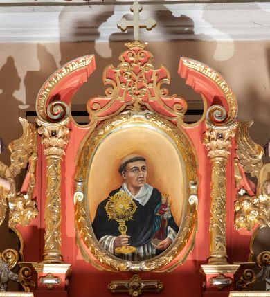 Zdjęcie nr 2: Obraz w kształcie stojącego owalu w złoconej ramie utworzonej z liści laurowych z rollwerkiem. W centrum ukazany w półpostaci św. Jacek, który w prawej ręce trzyma monstrancję a w lewej figurkę Matki Boskiej z Dzieciątkiem. Święty na owalną twarz, z ciemnymi, szeroko otwartymi oczami, wąski nos i niewielkie usta, na głowie wyciętą tonsurę. Jest ubrany w strój zakonny – biały habit i czarną pelerynę. Nad głową ma nimb. Tło obrazu jasne, ciemniejące za postacią świętego. 