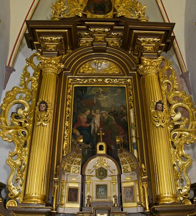 Zdjęcie nr 41: Ołtarz drewniany, architektoniczny, jednoosiowy, jednokondygnacyjny, ze zwieńczeniem, z dwiema bramkami. Jest umieszczony na murowanym, dwustopniowym podwyższeniu. Podstawa ołtarza murowana, skrzyniowa o kształcie prostopadłościanu, o obudowie drewnianej. Brak kamiennej mensy ołtarzowej. W partii antepedium nisza w kształcie prostokąta leżącego. Na mensie ustawione tabernakulum w formie centralnego kościółka zwieńczonego kopułą z latarnią.

Retabulum jest flankowane dwiema złoconymi kolumnami kompozytowymi, ustawionymi na profilowanych cokołach. Trzony kolumn są kanelowane, ozdobione w połowie wysokości główkami aniołków ujętymi dekoracją chrząstkową. Kolumny dźwigają belkowanie z wydatnym, przełamującym się nad nimi gzymsem, ozdobione ornamentem małżowinowym. W polu środkowym ołtarza profilowana, złocona i malowana rama z górną krawędzią w formie arkady z kluczem, dolna krawędź zaakcentowana uskokiem. W niej druga rama, prostokątna, ozdobiona ornamentem małżowinowym i złocona, z obrazem przedstawiającym Św. Annę Samotrzeć w otoczeniu świętych. We wnęce, za zasuwą, widnieje obraz Matki Boskiej z Dzieciątkiem w prostokątnej ramie, rzeźbionej i złoconej, pokrytej ornamentem roślinnym. Wnęka pokryta ciemnoczerwonym aksamitem, poniżej obrazu zawieszone wota. W polu arkady, w półkolistym tympanonie, na tle złoconych obłoków srebrna gołębica Ducha Świętego. Na gzymsie, na osi kolumn wazony z kwiatami. Po bokach retabulum bogato rzeźbione i złocone uszy z bujnym ornamentem małżowinowym. W zwieńczeniu, w owalnej, profilowanej i złoconej ramie obraz przedstawiający Wniebowzięcie Najświętszej Marii Panny. Rama otoczona szeroko rozbudowaną ornamentyką małżowinową. Po bokach ołtarza bramki prowadzące za ołtarz, o kształcie prostokąta stojącego, ze zwieńczeniem w formie łuku kotarowego zamkniętego od góry prostym odcinkiem, z profilowanym cokołem (pod figurę) z podwieszonym ornamentem w kształcie muszli. Górne narożniki bramek podkreślone złoconym ornamentem w kształcie wolut z elementem roślinnym. Otwór wejściowy, w kształcie analogicznym do bramki, z profilowanymi krawędziami, zakryty kotarą z aksamitu w kolorze ciemnoczerwonym. Struktura ołtarza polichromowana w kolorze ciemnobrązowym. Kolumny, pilastry, profilowania i dekoracja rzeźbiarska złocone.
