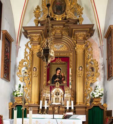 Zdjęcie nr 8: Ołtarz drewniany, architektoniczny, jednoosiowy, jednokondygnacyjny, ze zwieńczeniem, z dwiema bramkami. Jest umieszczony na murowanym, dwustopniowym podwyższeniu. Podstawa ołtarza murowana, skrzyniowa o kształcie prostopadłościanu, o obudowie drewnianej. Brak kamiennej mensy ołtarzowej. W partii antepedium nisza w kształcie prostokąta leżącego. Na mensie ustawione tabernakulum w formie centralnego kościółka zwieńczonego kopułą z latarnią.

Retabulum jest flankowane dwiema złoconymi kolumnami kompozytowymi, ustawionymi na profilowanych cokołach. Trzony kolumn są kanelowane, ozdobione w połowie wysokości główkami aniołków ujętymi dekoracją chrząstkową. Kolumny dźwigają belkowanie z wydatnym, przełamującym się nad nimi gzymsem, ozdobione ornamentem małżowinowym. W polu środkowym ołtarza profilowana, złocona i malowana rama z górną krawędzią w formie arkady z kluczem, dolna krawędź zaakcentowana uskokiem. W niej druga rama, prostokątna, ozdobiona ornamentem małżowinowym i złocona, z obrazem przedstawiającym Św. Annę Samotrzeć w otoczeniu świętych. We wnęce, za zasuwą, widnieje obraz Matki Boskiej z Dzieciątkiem w prostokątnej ramie, rzeźbionej i złoconej, pokrytej ornamentem roślinnym. Wnęka pokryta ciemnoczerwonym aksamitem, poniżej obrazu zawieszone wota. W polu arkady, w półkolistym tympanonie, na tle złoconych obłoków srebrna gołębica Ducha Świętego. Na gzymsie, na osi kolumn wazony z kwiatami. Po bokach retabulum bogato rzeźbione i złocone uszy z bujnym ornamentem małżowinowym. W zwieńczeniu, w owalnej, profilowanej i złoconej ramie obraz przedstawiający Wniebowzięcie Najświętszej Marii Panny. Rama otoczona szeroko rozbudowaną ornamentyką małżowinową. Po bokach ołtarza bramki prowadzące za ołtarz, o kształcie prostokąta stojącego, ze zwieńczeniem w formie łuku kotarowego zamkniętego od góry prostym odcinkiem, z profilowanym cokołem (pod figurę) z podwieszonym ornamentem w kształcie muszli. Górne narożniki bramek podkreślone złoconym ornamentem w kształcie wolut z elementem roślinnym. Otwór wejściowy, w kształcie analogicznym do bramki, z profilowanymi krawędziami, zakryty kotarą z aksamitu w kolorze ciemnoczerwonym. Struktura ołtarza polichromowana w kolorze ciemnobrązowym. Kolumny, pilastry, profilowania i dekoracja rzeźbiarska złocone.
